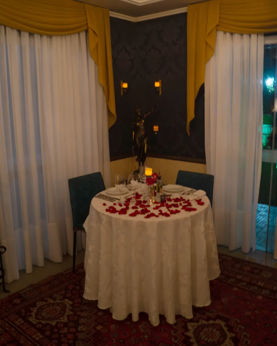 Mesa para dois no salão do Cordon Blanc, decorada com pétalas de rosa e vela, entre cortinas douradas